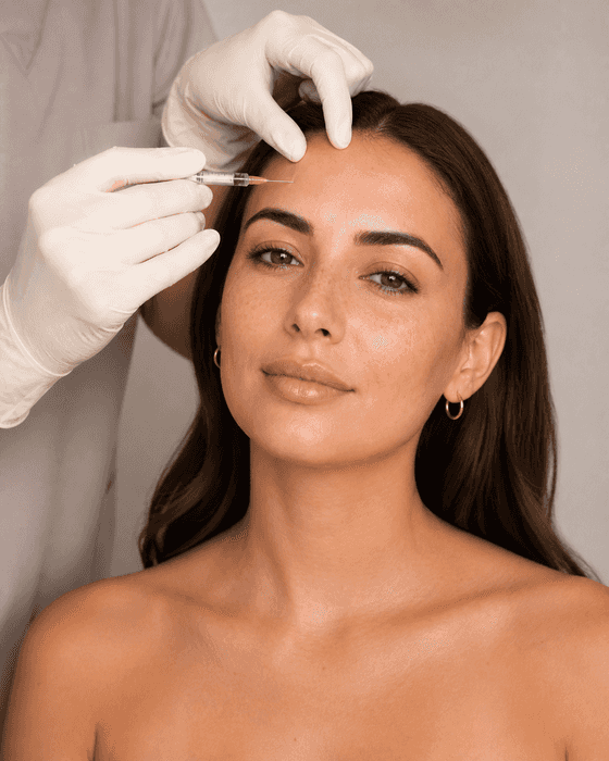 A clinician in cream gloves steadies a client’s forehead and holds a small clear syringe to the skin above her outer brow.