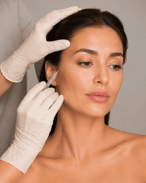 A clinician in cream gloves rests one hand at a client’s hairline and angles a clear syringe up toward her temple.