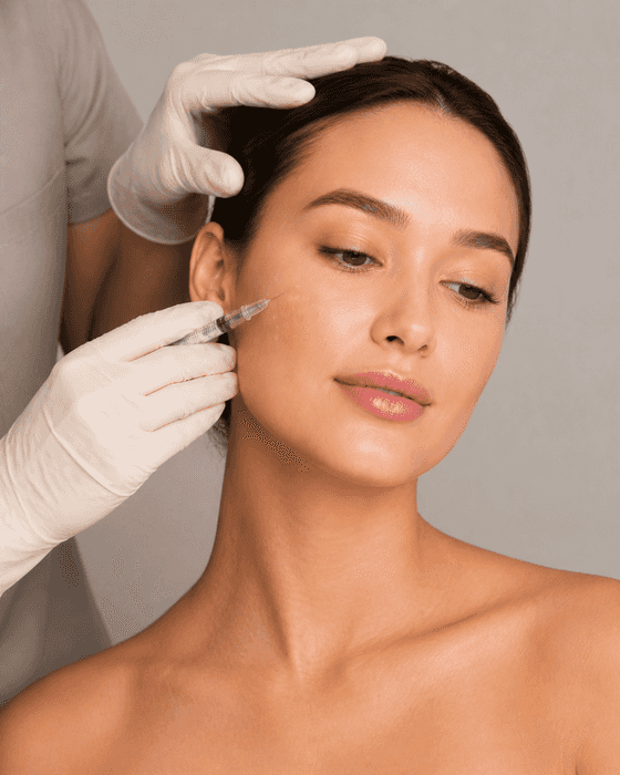 A clinician in cream gloves steadies a client’s hairline and holds a small clear syringe horizontally to her upper cheek.
